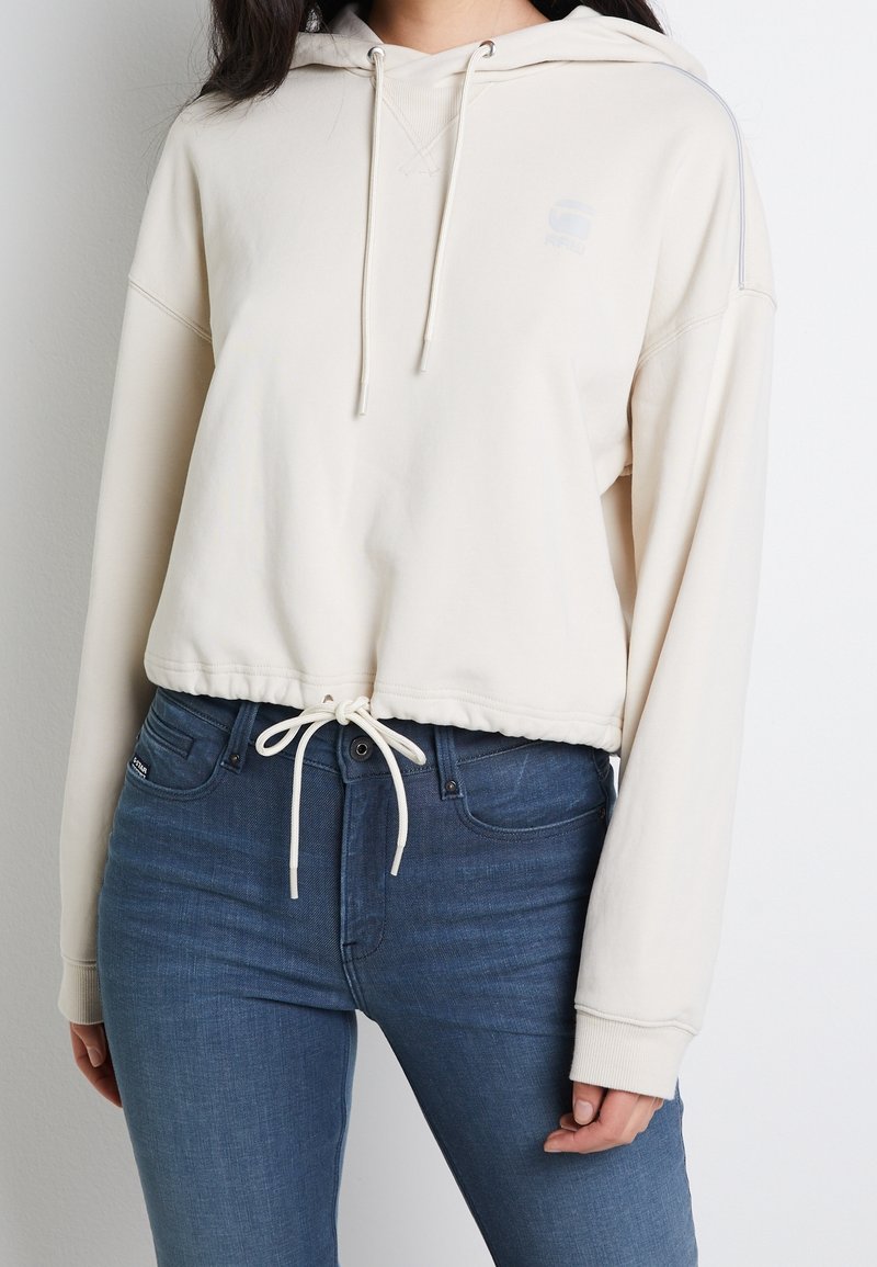 Krämfärgad kort hoodie med dragsko och ribbade ärmslut, med en logodetalj på bröstet och en avslappnad passform. Matchad med blå jeans.