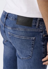 Sininen farkkuhousut, joissa on takana musta nahkapaikka, jossa lukee "ONLY & SONS". Housuissa on tavalliset taskut ja haalistunut rakenne.