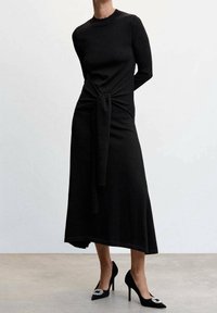 Robe noire en tricot avec manches longues, détail de taille nouée et ourlet fluide. Associée à des talons noirs à bout pointu ornés d'un accent décoratif.