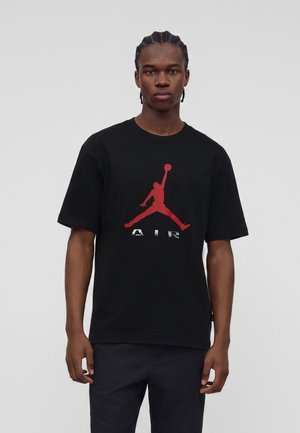 Ung mand med flettet hår iført en sort T-shirt med et stort rødt Air Jordan-logo og sorte bukser, står mod en ensfarvet baggrund.