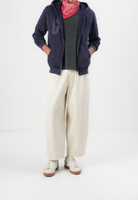Persona che indossa un felpa blu navy, una maglietta verde scuro, un bandana rossa, pantaloni larghi beige e sneakers bianche, in piedi con le mani nelle tasche.