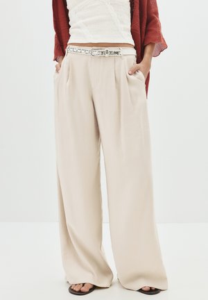 Pantalon large beige avec des plis, doté de poches latérales et d'une ceinture studded argentée. Tissu doux avec une texture lisse.
