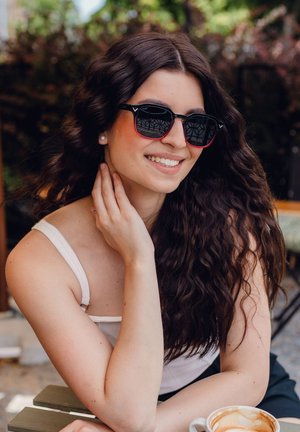 Black sunglasses with a red gradient frame, featuring square lenses and a sleek design. Long wavy hair and a light tank top are visible.