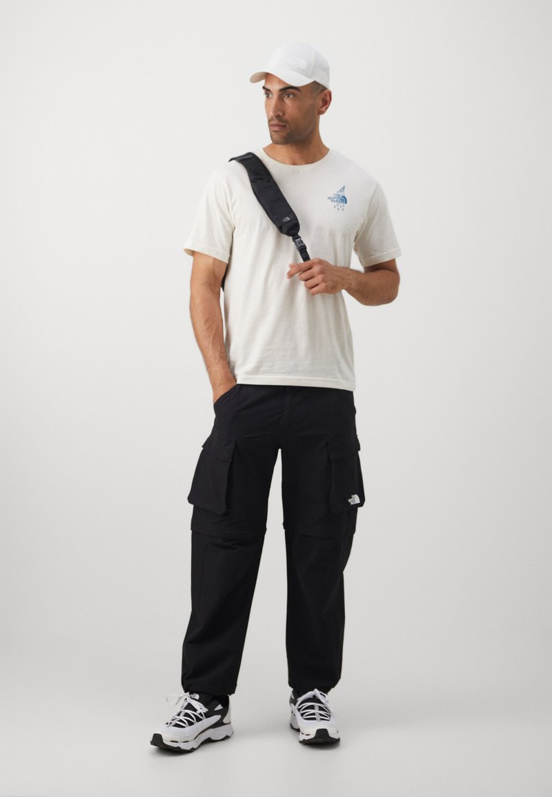 Hvit caps, beige kortermet T-skjorte med blå grafikk, svarte cargo-bukser med lommer, og svart-og-hvite joggesko. Uformell antrekk.