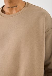 Nakketatovering med ordene "tro på deg selv" på en person som har på seg en beige genser mot en lys bakgrunn.