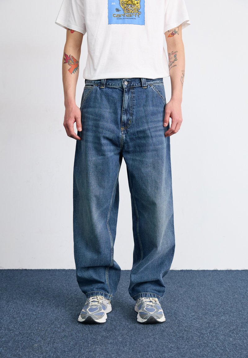 Vaqueros de mezclilla azul holgados con parches desgastados y una cintura alta, combinados con una camiseta gráfica blanca y zapatillas con estampado.