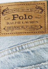 Brown leather tag with embossed text "Polo Ralph Lauren," stitched onto light blue denim with a faded texture and yellow thread.