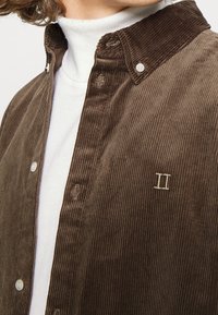 Camisa de pana marrón con cuello abotonado, que presenta líneas verticales texturizadas, botones de perla y un pequeño detalle bordado de "II".