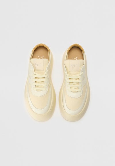Cream-colored sneakers with a smooth fabric upper, textured accents, and a thick, beige rubber sole. Features round laces and subtle logo.