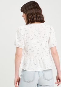 Blouse blanche à fleurs avec des manches bouffantes et un ourlet peplum, présentant de petits motifs floraux noirs et un tissu texturé. Portée avec un jean en denim clair.