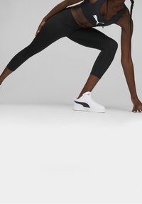 Czarne legginsy sportowe o dopasowanym kroju i skróconej długości, zestawione z białymi sneakersami z czarnym logo w kształcie łyżwy. Gładka tekstura.