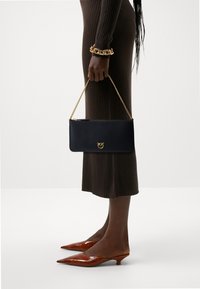 Pochette en cuir noir avec une poignée en chaîne dorée, arborant un emblème doré. Modèle portant une robe marron à côtes et des talons aiguilles marron.