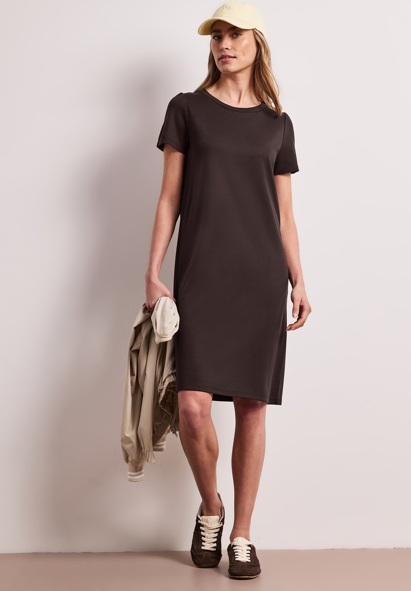 Femme portant une robe marron foncé au genou, une casquette beige, tenant une veste beige et portant des baskets marron, debout devant un mur clair uni.