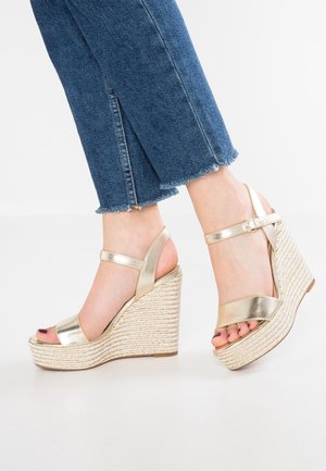 Złote metaliczne sandały na koturnie z plecioną platformą z juty, z paskiem na kostkę i otwartym noskiem. Stylizacja z postrzępionymi dżinsowymi spodniami.