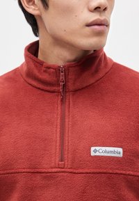Homme portant un pull polaire rouge avec une fermeture éclair au quart et un logo Columbia visible sur la poitrine, légèrement tourné vers la droite.