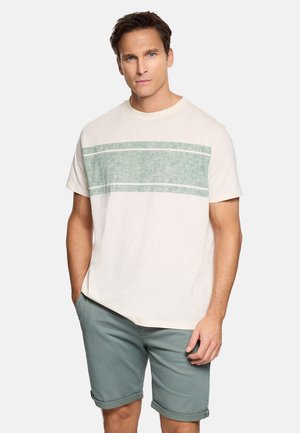 Hombre con camiseta beige claro con una franja horizontal verde y pantalones cortos verde apagado remangados, de pie con una mano en el bolsillo.