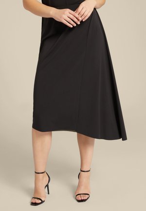 Zwarte asymmetrische rok van gladde stof, met een diagonale onderzoom. Een paar zwarte strappy hakken maakt de look compleet.
