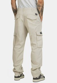 Beige cargobroek met zijzakken, elastische tailleband en een losse pasvorm. Heeft zwarte merkemblemen en wordt gedragen met zwart-witte sneakers.