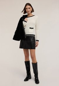 Donna che indossa un maglione bianco con texture e bordi neri, una minigonna in pelle nera, stivali neri fino al ginocchio, mentre tiene una giacca di pelliccia nera su una spalla.