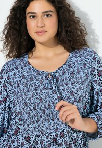 Niebieska kwiatowa bluzka z wiązaniem przy dekolcie, marszczoną fakturą oraz wzorami w kolorach czarnym, czerwonym i białym. Długie rękawy o luźnym kroju.