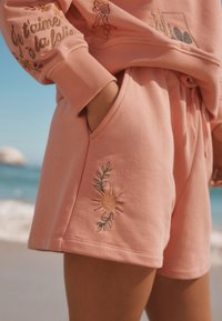 Rosa Baumwollshorts mit gesticktem Sonnen- und Blattemuster. Der Bund ist elastisch mit einem Kordelzug. Außeneinstellung am Strand.