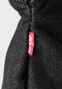 Κόκκινη ετικέτα ύφασματος Levi's σε μαύρο τζιν με εμφανείς λευκές ραφές και υφή υφαντής.