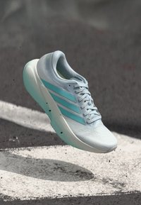 Svetlo modna športna obutev s teksturirano površino, tremi zelenkastimi črtami in zaobljeno podplatom. Ima vezalko in oblazinjen petni del.