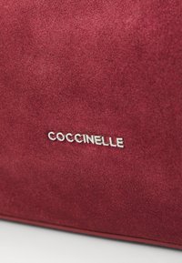 Deep burgundy suede bag featuring a metallic silver "Coccinelle" logo on a smooth surface. Elegant and stylish accessory focus.