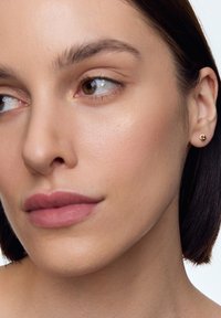 Złote kolczyki wkrętki z kwiatowym wzorem, o gładkiej powierzchni i małym, okrągłym kształcie, noszone na neutralnym odcieniu skóry.