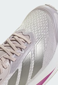 Svetlosivá športová obuv s texturovaným sieťovaným zvrškom, striebornými akcentovými pruhmi a bledoružovou podrážkou. Má ploché šnúrky a vankúšikový dizajn.