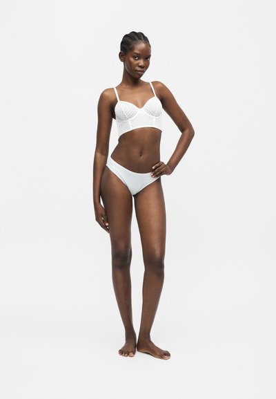 Λεπτή γυναίκα στέκεται ξυπόλυτη με το χέρι στη μέση, φοράει λευκό δαντελένιο bralette και ταιριαστό εσώρουχο μπικίνι, σε μονόχρωμο φόντο.