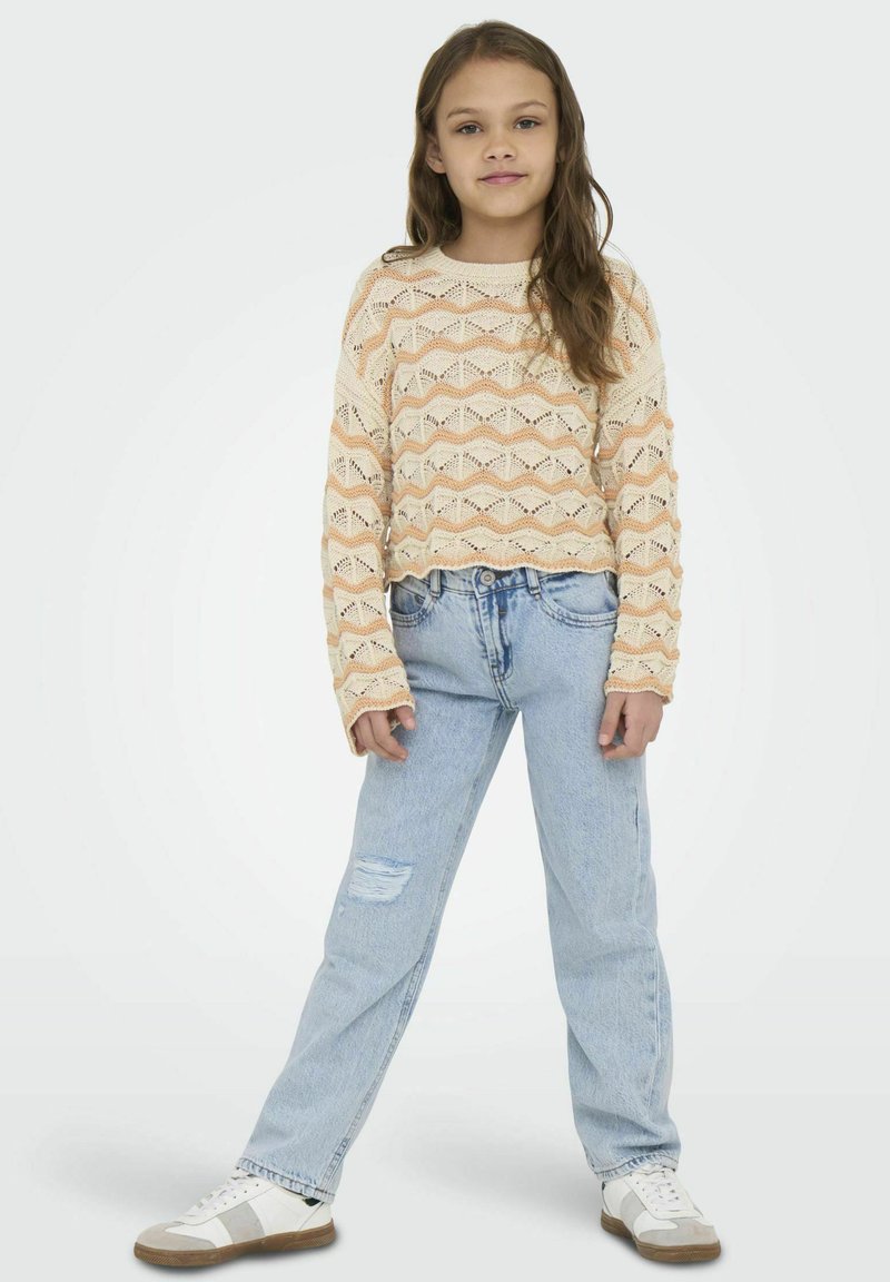 Gebreide sweater in licht perzik en crème met zigzagpatroon, gecombineerd met lichtblauwe jeans met een scheurdetail en witte sneakers.