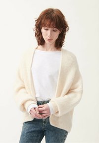 Fluffy cream cardigan with open front, batwing sleeves, and ribbed texture, worn over a white shirt and blue jeans.