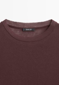 Burgunderfarbendes T-Shirt mit Rundhalsausschnitt aus Baumwolle. Es hat eine weiche Textur und einen genähten Halsausschnitt mit einem Etikett, das "Massimo Dutti, Made in Portugal" trägt.