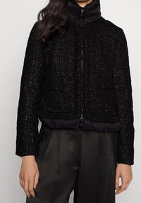 Black, cropped jacket combines textured woven fabric with a puffy, quilted black collar and bottom hem. Features a central zipper.