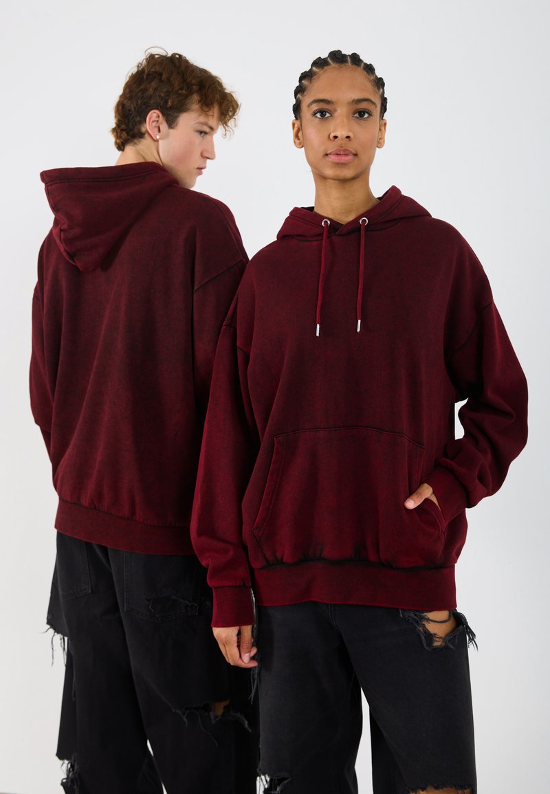 Burgunderfarbene Kapuzenjacke mit einer Vordertasche und Zugbändern, kombiniert mit verwaschenen schwarzen Hosen. Weicher Stoff und lässiger Schnitt.