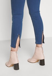 Jeans ajustados azules con cremalleras en los tobillos, combinados con botines de tacón en un suave tono rosa claro, que cuentan con una cremallera en la parte trasera y un tacón negro grueso.