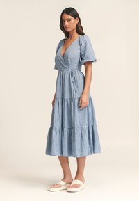 Sininen gingham-kuvioinen kietaisumekko, jossa lyhyet pallohihat, kerroksittainen helma ja säädettävä vyötärönauha. Mekon kanssa on vaaleat korolliset sandaalit.