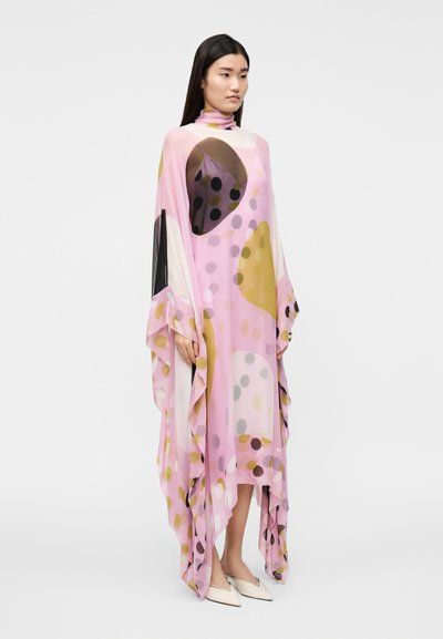 Žena v splývavej ružovej sukni s veľkými abstraktnými kruhmi, stojaca na bielom pozadí, v kombinácii s bielymi špicatými topánkami.