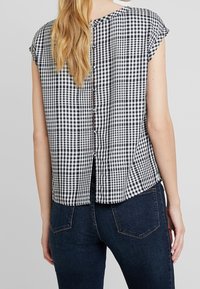 Zadní pohled na zkrácený top s krátkým rukávem, který má černo-bílý kostkovaný vzor, zaoblený spodní lem a zapínání na knoflíky ve středu.