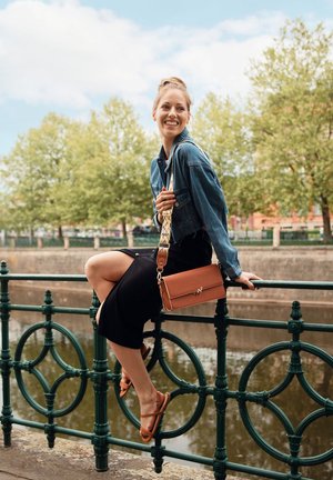 Tan leather handbag with gold hardware and a patterned strap, accompanied by a denim jacket and black skirt. The setting has green railings and trees.