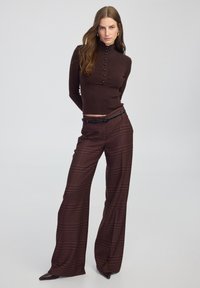 Braunfarbener hochgeschlossenes Langarmoberteil mit Knopfdetails, kombiniert mit weit geschnittenen Karohosen in Maroon und Braun mit einem schwarzen Gürtel.