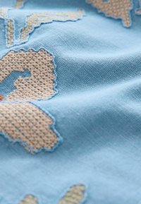Helles blaues Gewebe mit eingewebtem beigen Blumenmuster. Weiche Textur, leichter Glanz und detaillierte Stickerei an den Rändern.
