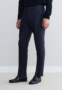 Prilagojene temno sive hlače iz teksturirane tkanine, s ploskim sprednjim delom, potisnjenimi šivi in zapiranjem z enim gumbom. Kombinirane s črnimi loaferji.