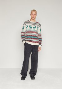 Gebreide trui in crème met groene rendieren en rode patroonaccenten, gecombineerd met zwarte losse broeken en zwarte sneakers. Glad textuur.