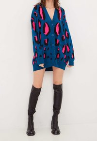 Modrý svetr s růžovými a černými leopardími skvrnami, s hlubokým výstřihem do V, knoflíkovým zapínáním a oversized střihem, doplněný černými vysokými kozačkami.