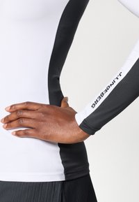 Pikad varrukad, sporditop, valge keha, mustad külgpaneelid ja "J.Lindeberg" tekst varrukal. Valmistatud siledast ja venivast materjalist.