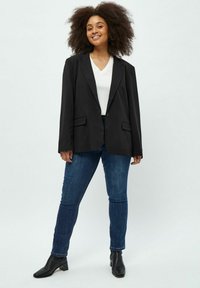 Blazer nero oversized con un solo bottone, due tasche anteriori, indossato sopra una t-shirt bianca con scollo a V, abbinato a jeans skinny scuri e stivali neri.