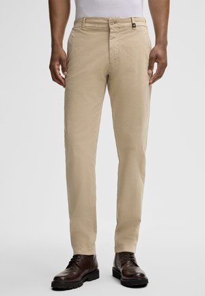 Beige chinos made of lightweight fabric. Features a flat front, two side pockets, and a button closure. Worn with brown leather boots.