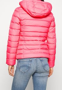 Polstrovaná růžová bunda s kapucí, s horizontálním prošíváním a hladkým povrchem. Nošená přes světle modré džíny s zadními kapsami.